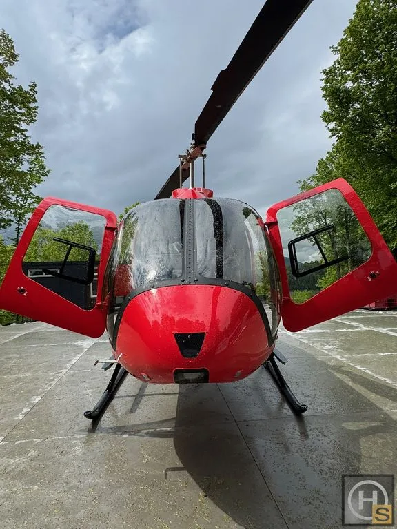 Front view of a red helicopter with its doors open, parked on a wet concrete surface outdoors.