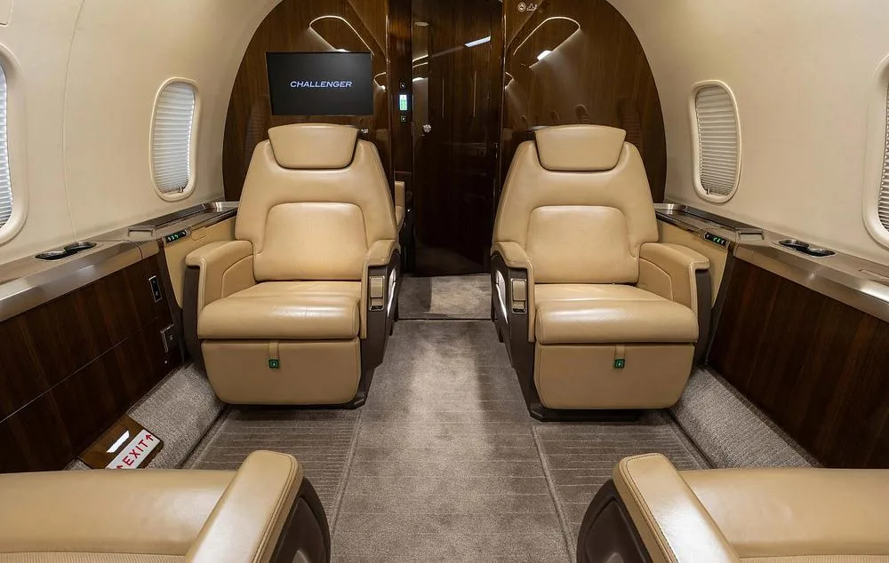 The luxurious interior of a Challenger business jet cabin with two beige leather seats and dark wood paneling.