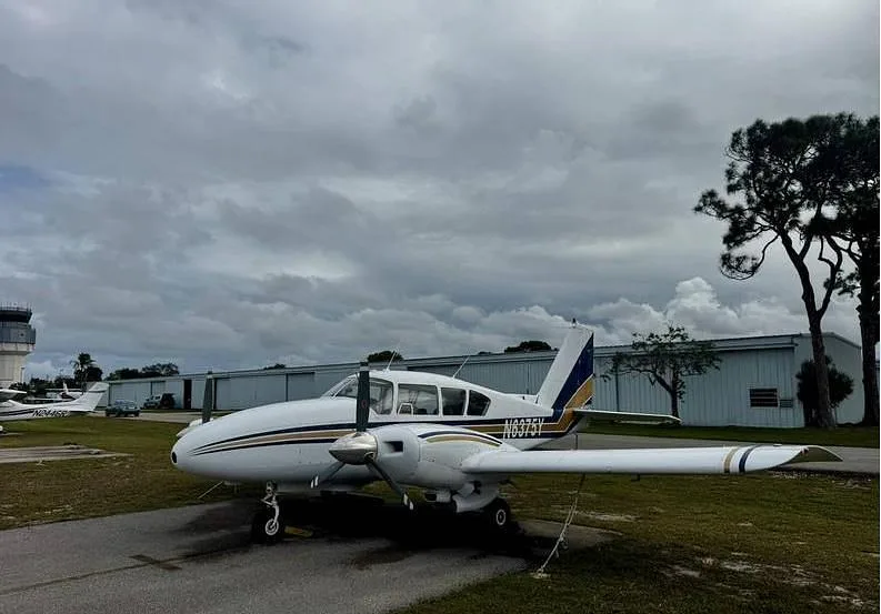 A side view of the white twin-engine aircraft N6375Y with blue and gold stripes, parked on an airfield with a control tower in the distance.