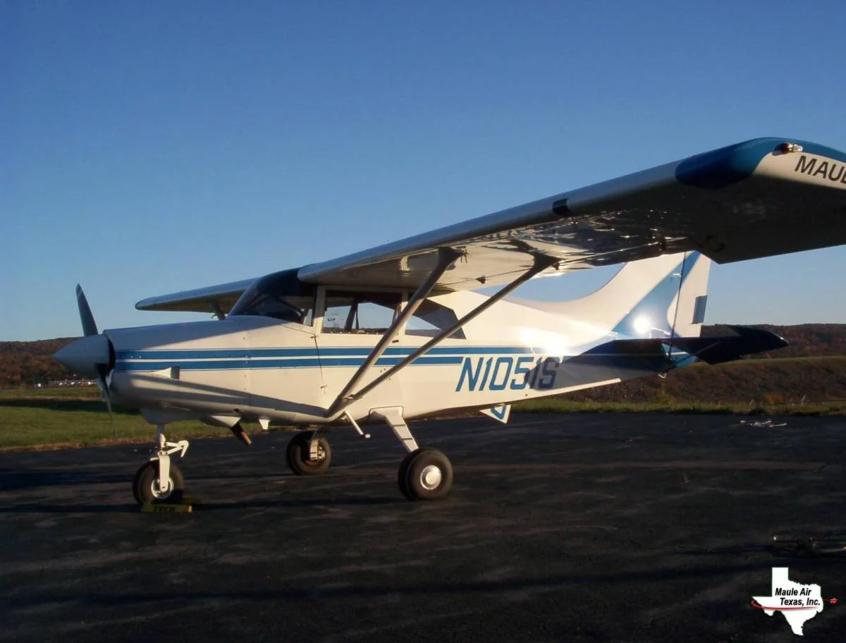 A white Maule aircraft with blue stripes is parked on a tarmac with hills in the background under a clear sky.
