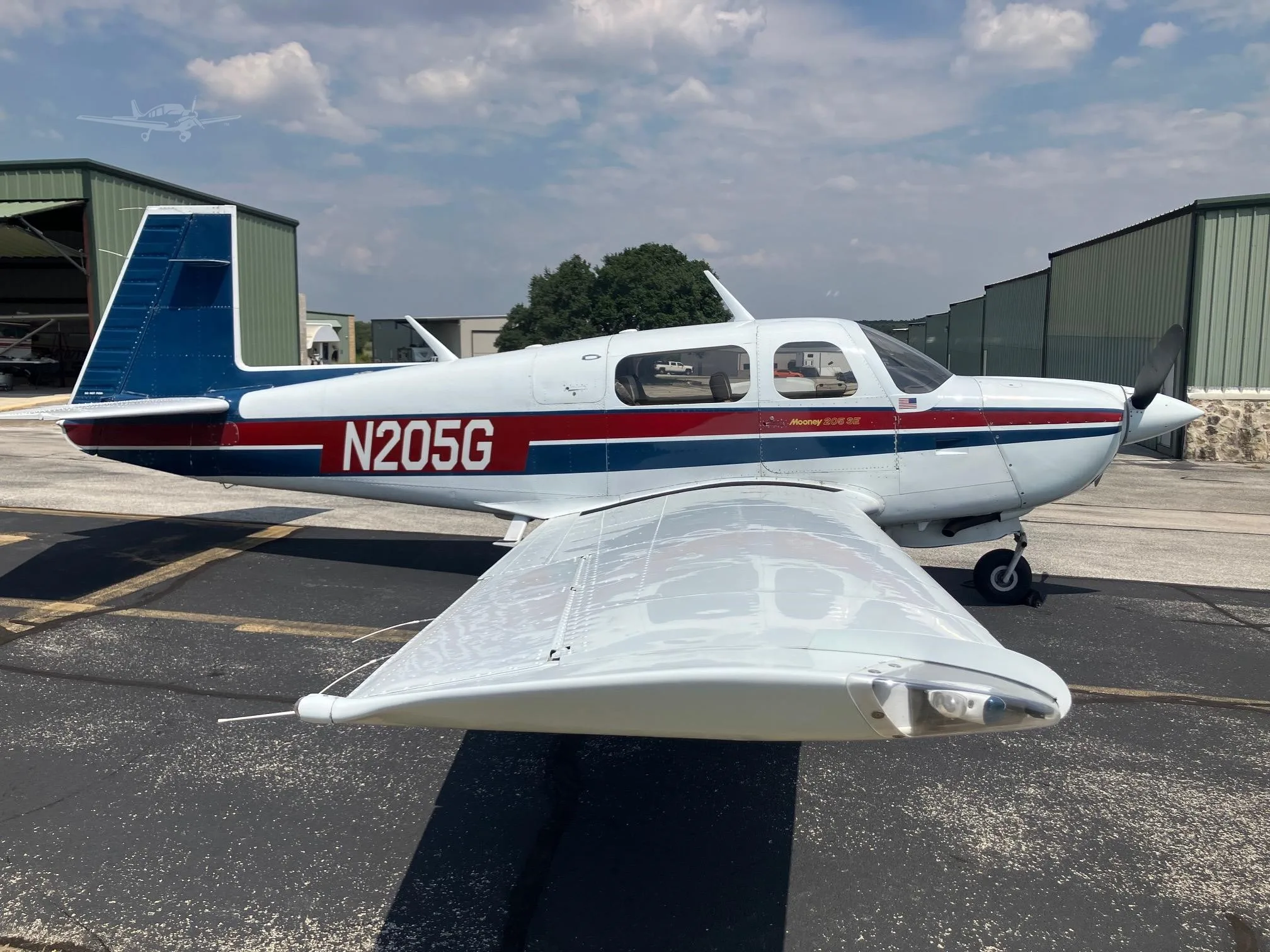 A white Mooney 205 SE aircraft with red and blue stripes is parked on an asphalt ramp with hangars in the background.