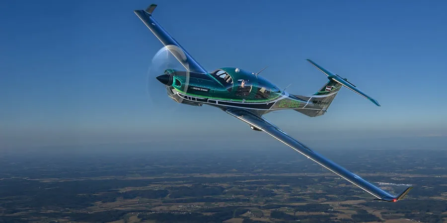 A green and black Diamond DA50 RG aircraft with 'DARK SHARK' livery flies against a blue sky over a forested landscape.