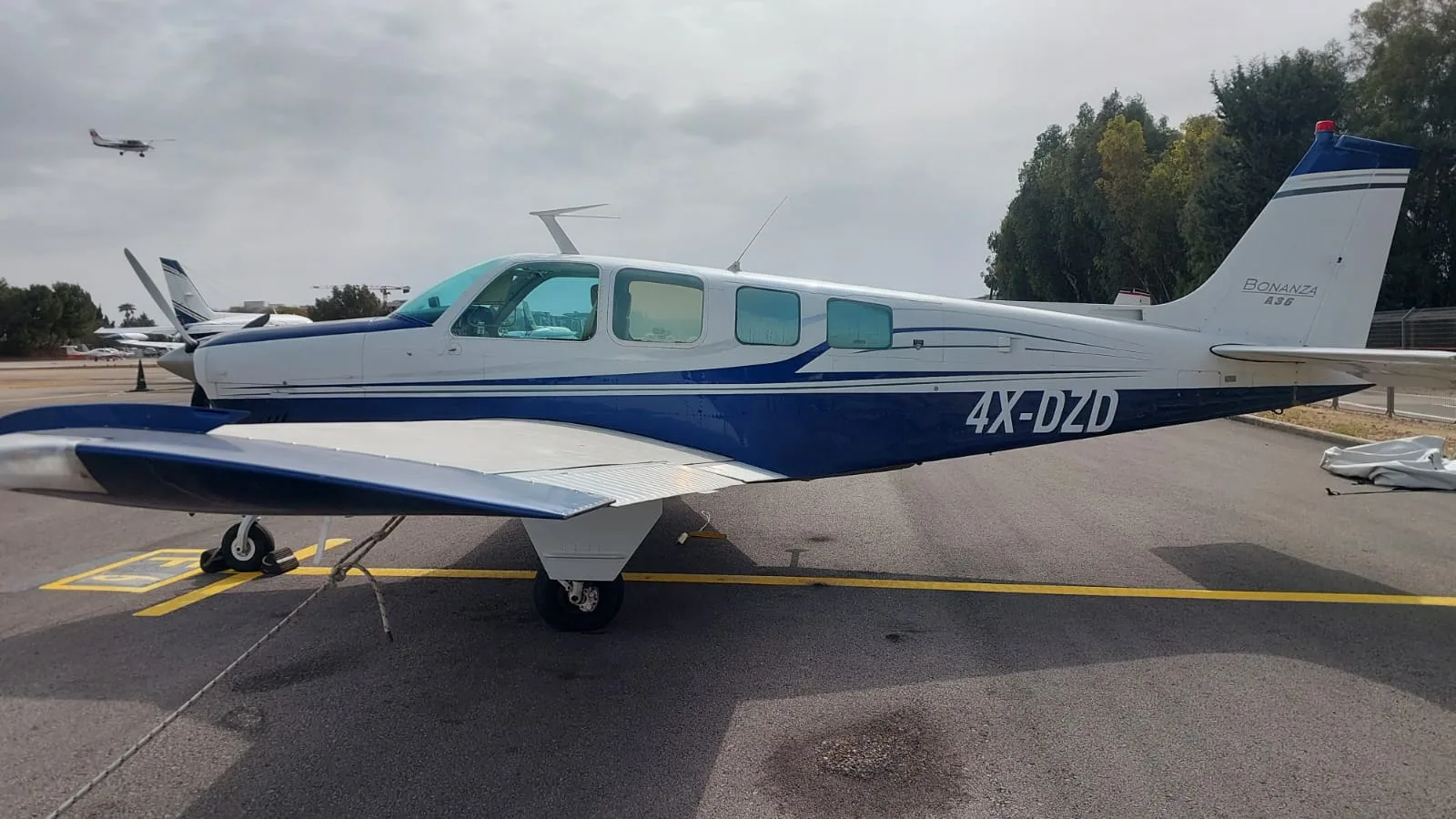 A white and blue Beechcraft Bonanza A36 aircraft with registration 4X-DZD parked on a tarmac at an airfield.