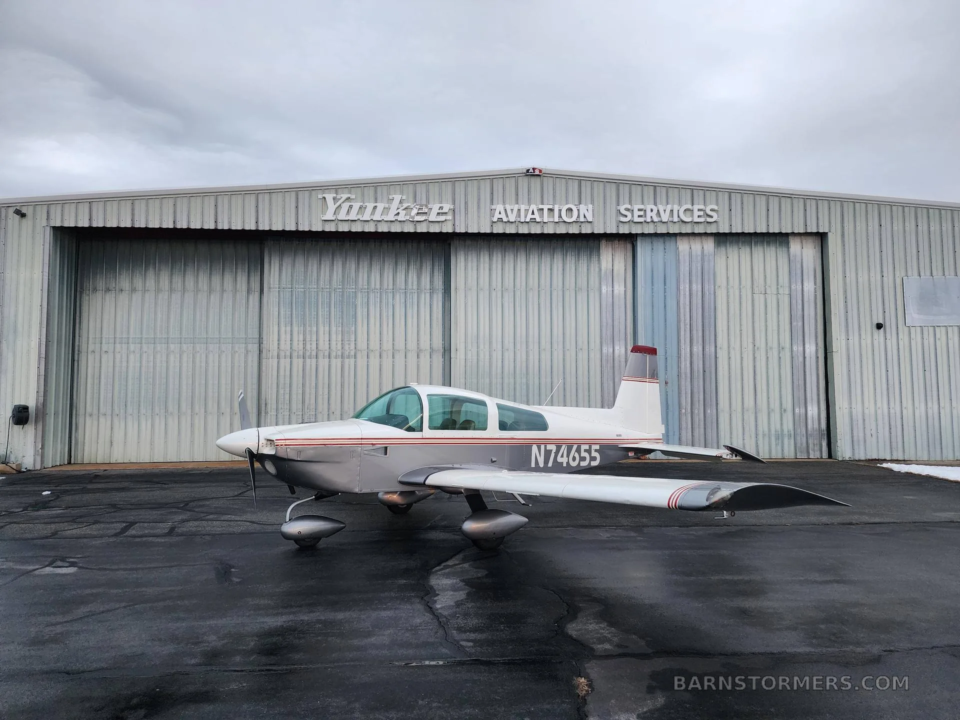 A white and silver Grumman AA-5 Traveler aircraft with red stripes parked on a wet tarmac in front of a hangar.