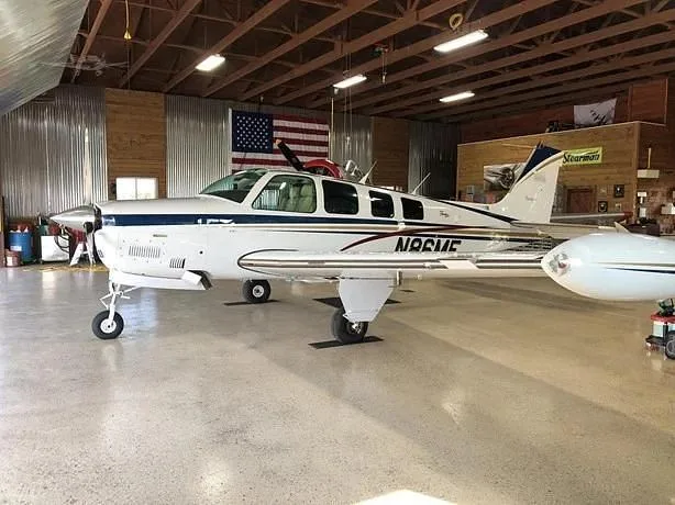 A white and blue Beechcraft Baron 58, registration N86ME, is parked in a hangar with an American flag in the background.