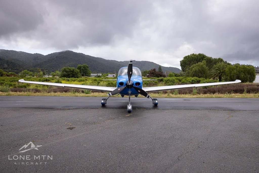 A front view of a blue and silver single-engine propeller aircraft (Cirrus SR22) with registration N248LF parked on a wet tarmac under a cloudy sky.