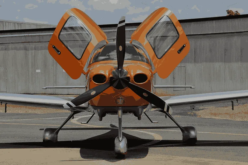 A front view of an orange and silver single-engine propeller aircraft with its gull-wing doors open on a tarmac.