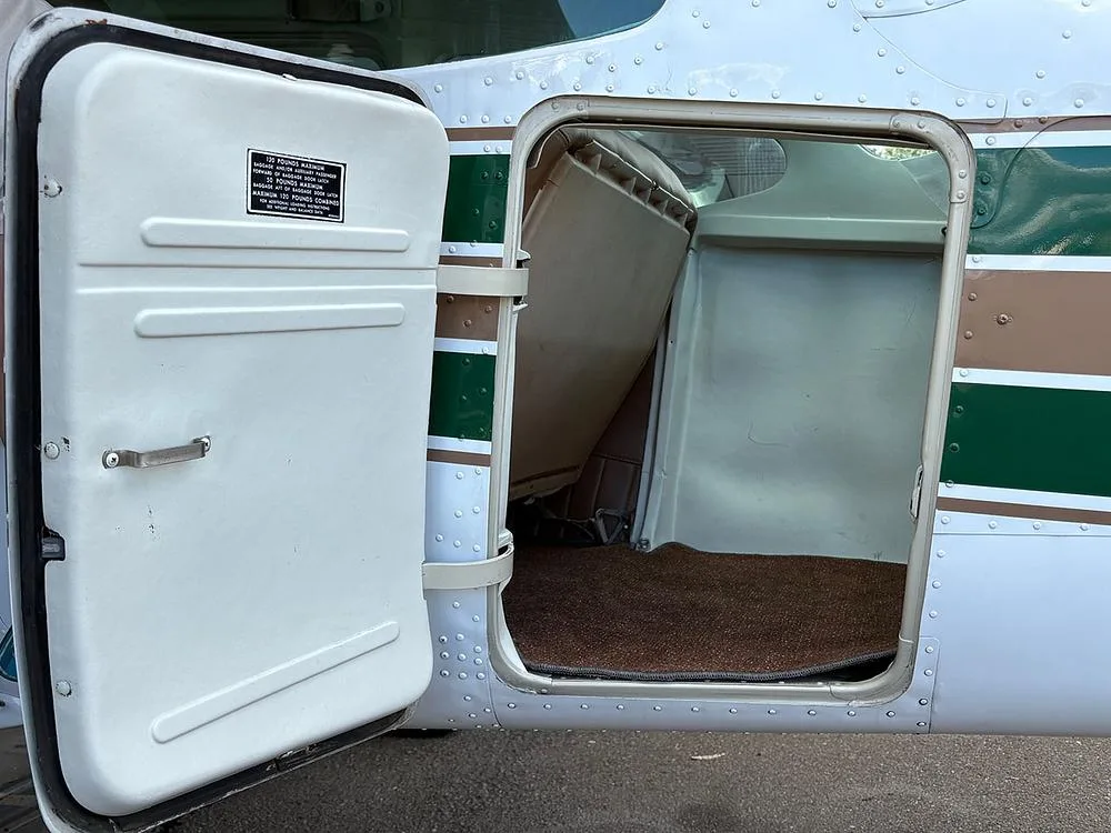 The open baggage compartment of the aircraft, showing a brown carpeted floor and a weight limit placard on the door.