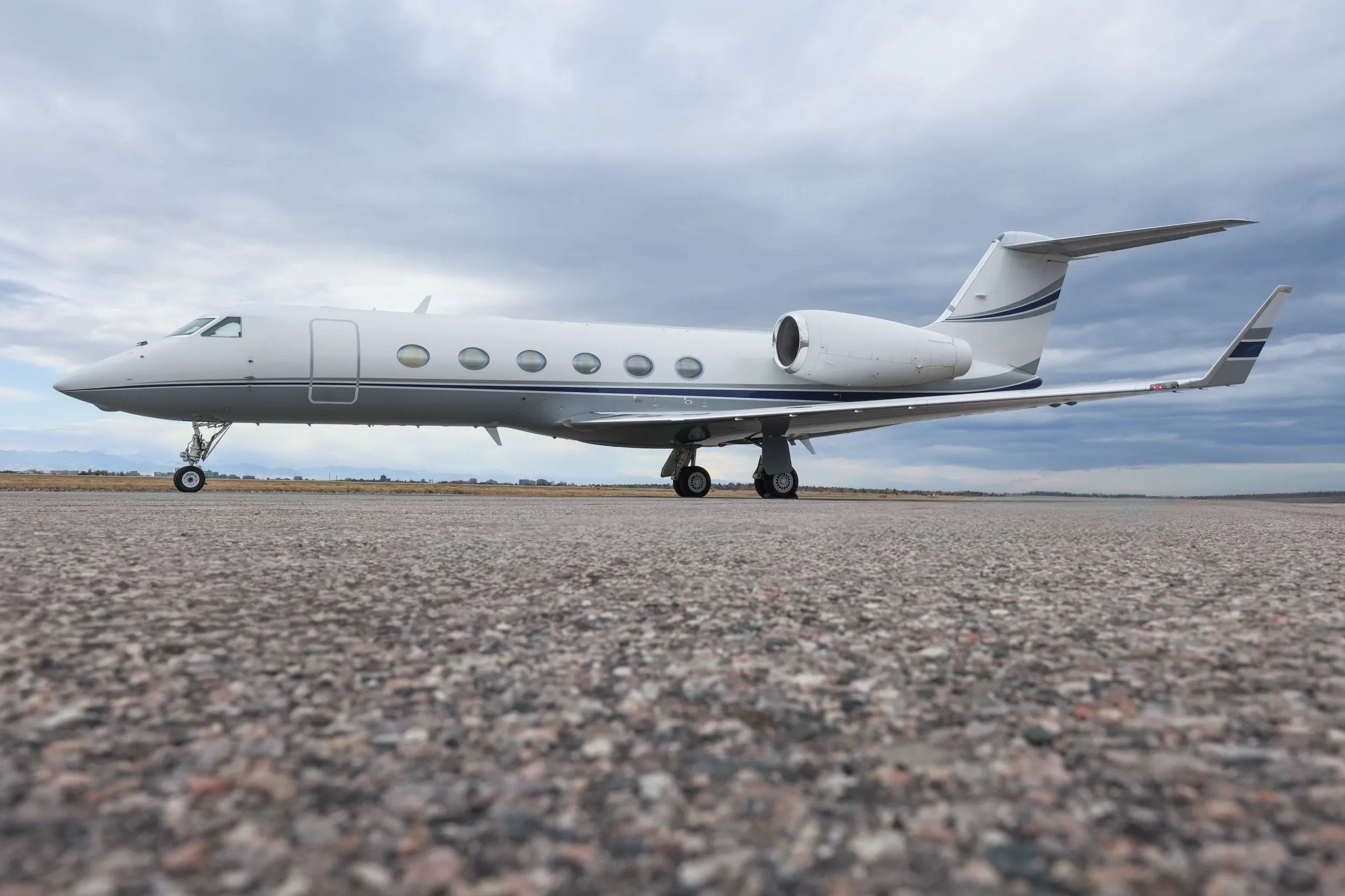 A white Gulfstream G550 business jet with blue accents on the tail and engines, parked on an asphalt runway under a cloudy sky.