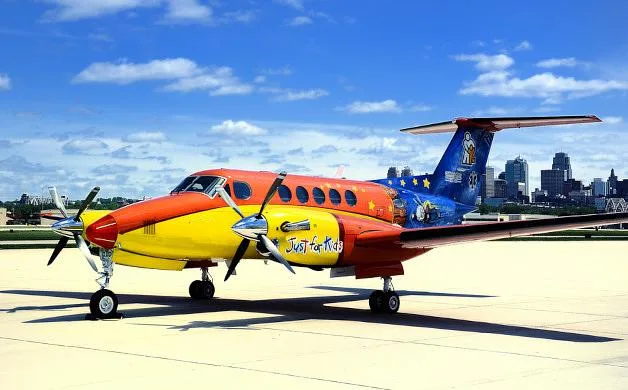 A King Air 350 aircraft with a vibrant orange, yellow, and blue 'Just for Kids' paint scheme, parked on a tarmac with a city skyline in the background.