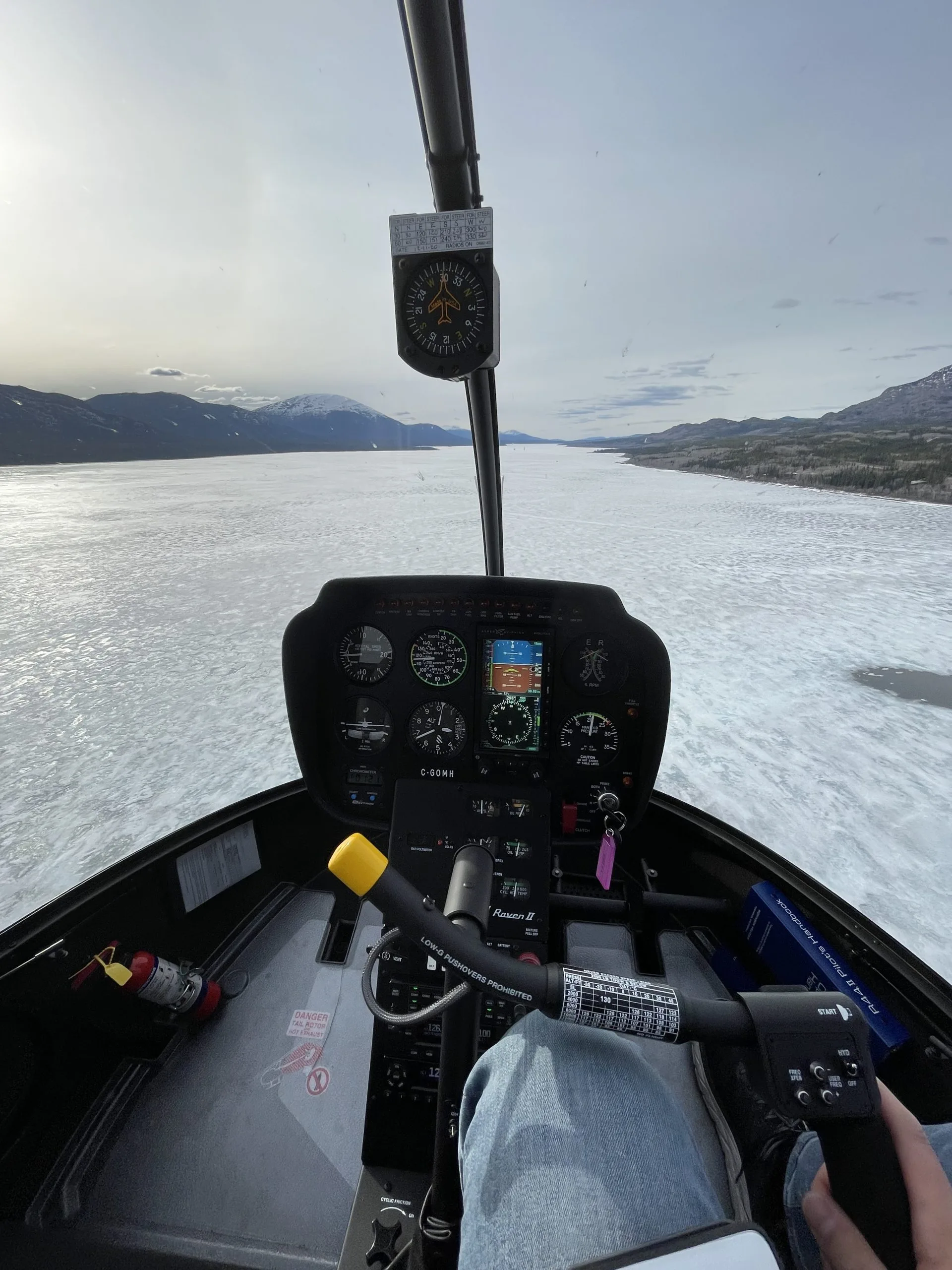 A pilot's view from the cockpit of a Robinson R44 Raven II helicopter, showing the instrument panel and a frozen lake surrounded by mountains.