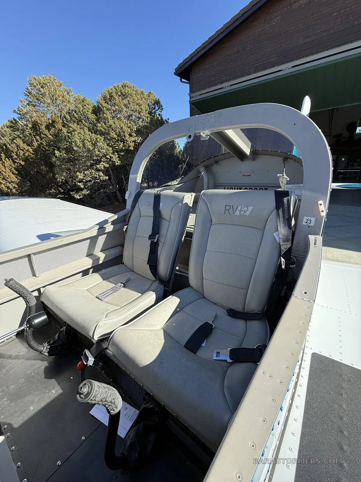 The light gray two-seat interior of an RV-12 aircraft is visible with seatbelts buckled.