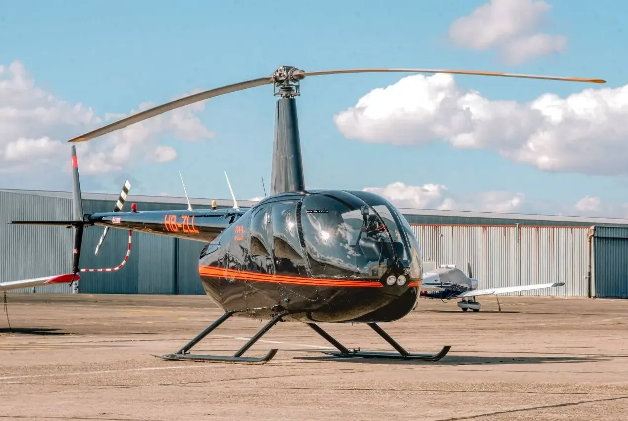 A black Robinson R44 Raven II helicopter, registration HB-ZLL, parked on an airfield with hangars in the background.