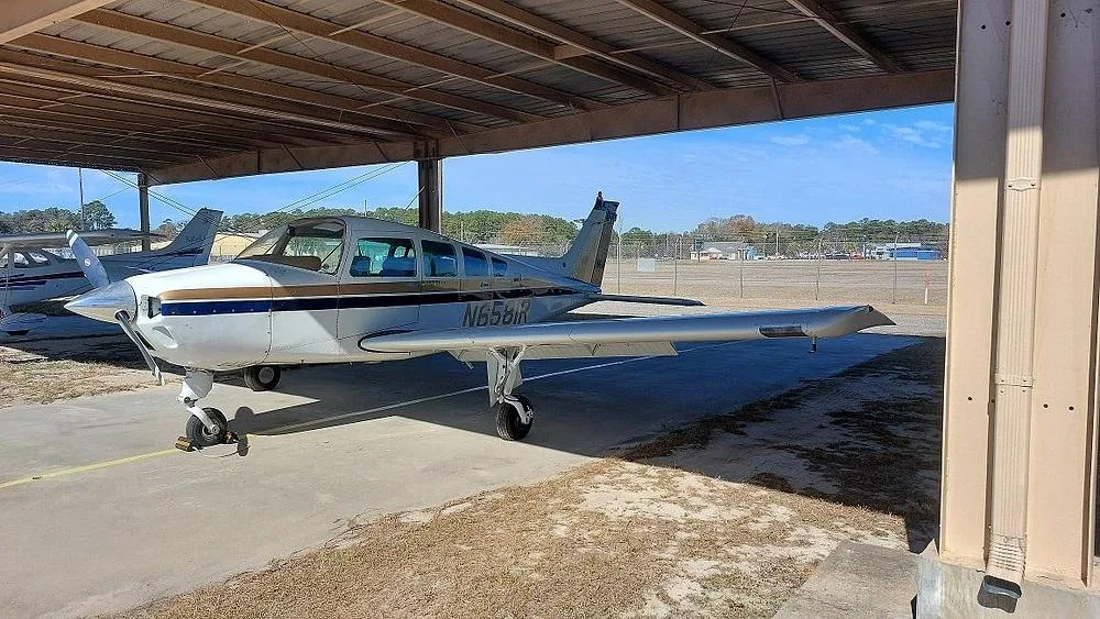A white and gold single-engine propeller aircraft (Cessna 177 Cardinal) with registration N6581R is parked under an open-sided hangar, viewed from the front-left.