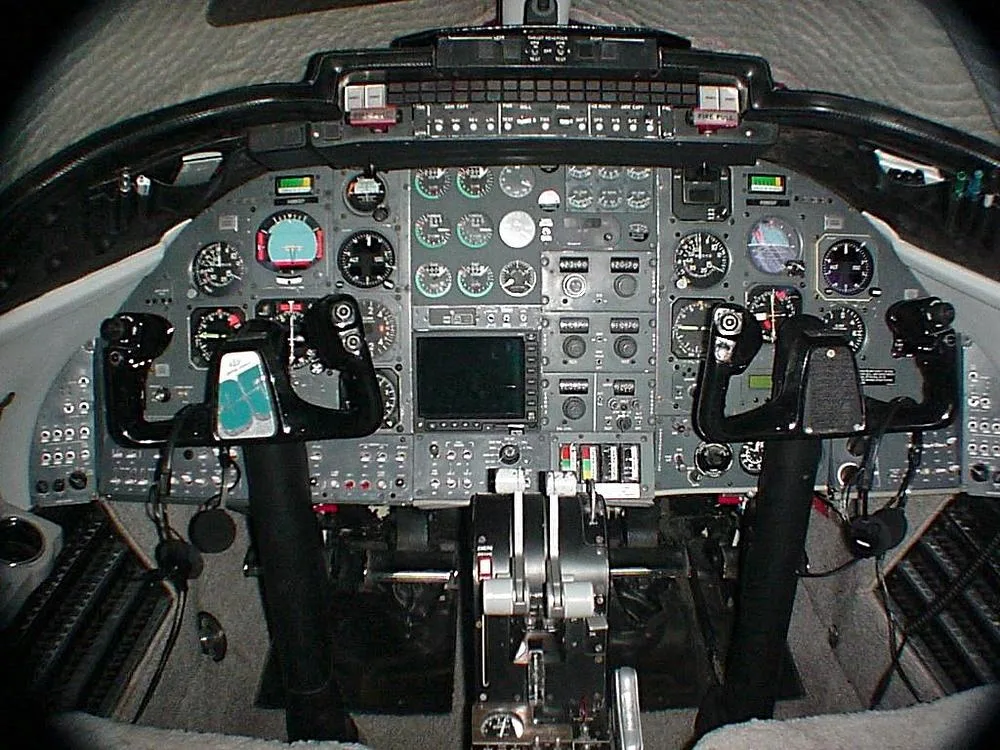 The cockpit of an aircraft features a complex instrument panel with multiple gauges, two control yokes, and throttle levers.