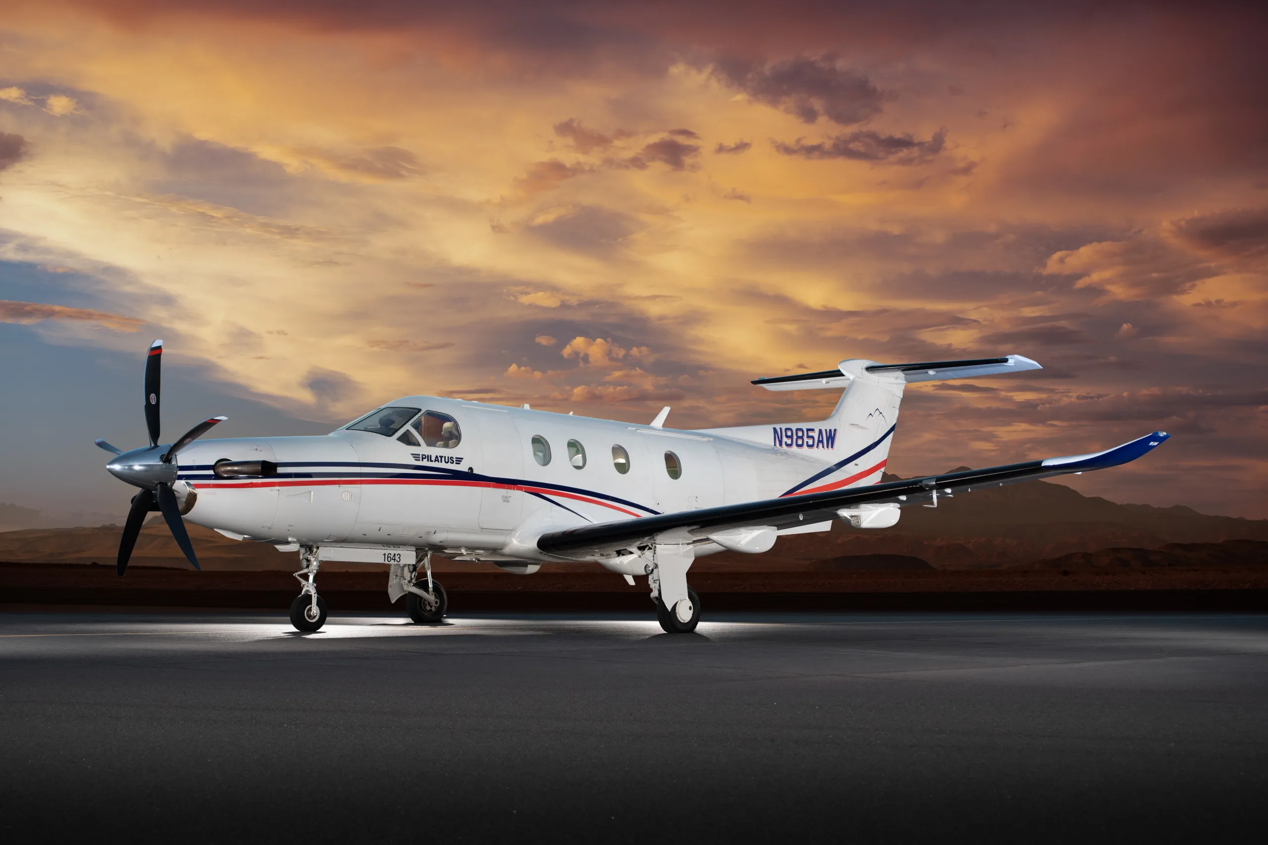 A Pilatus PC-12 aircraft, N985AW, with a white, blue, and red paint scheme, sits on a tarmac at sunset.