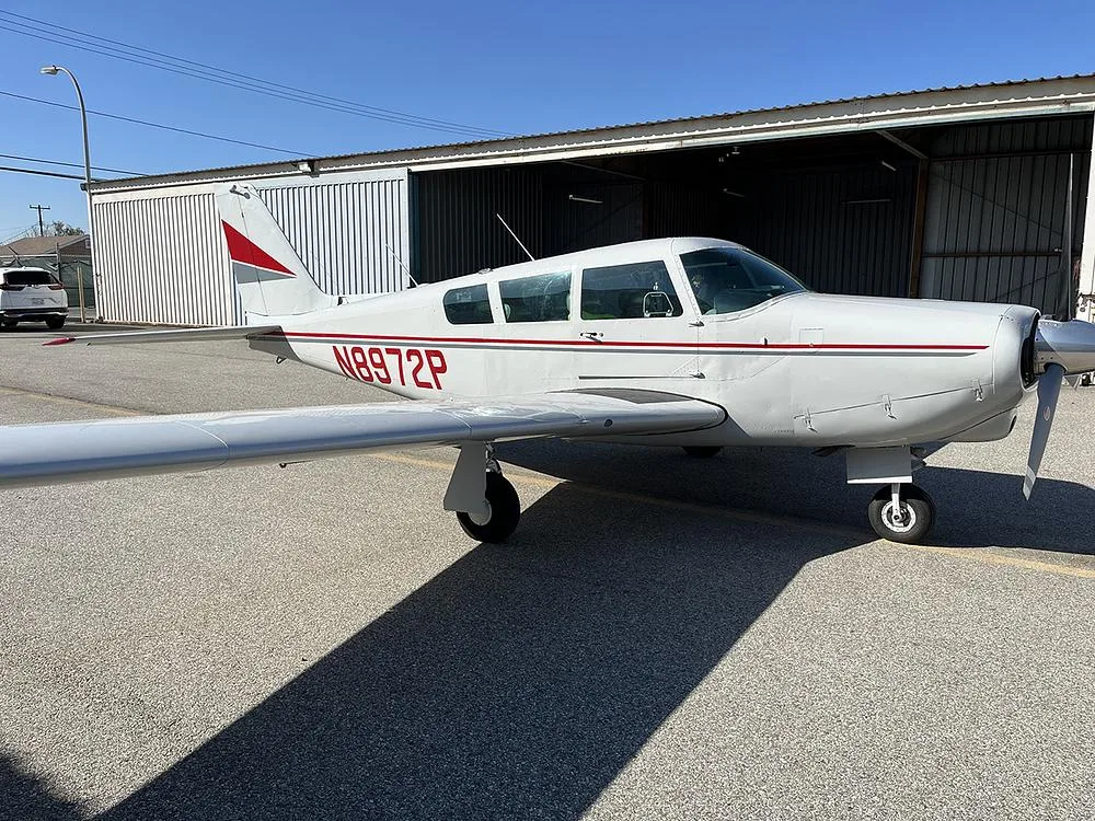 A white Piper Cherokee 180 (N8972P) with red and grey stripes parked on an asphalt ramp next to an open hangar.