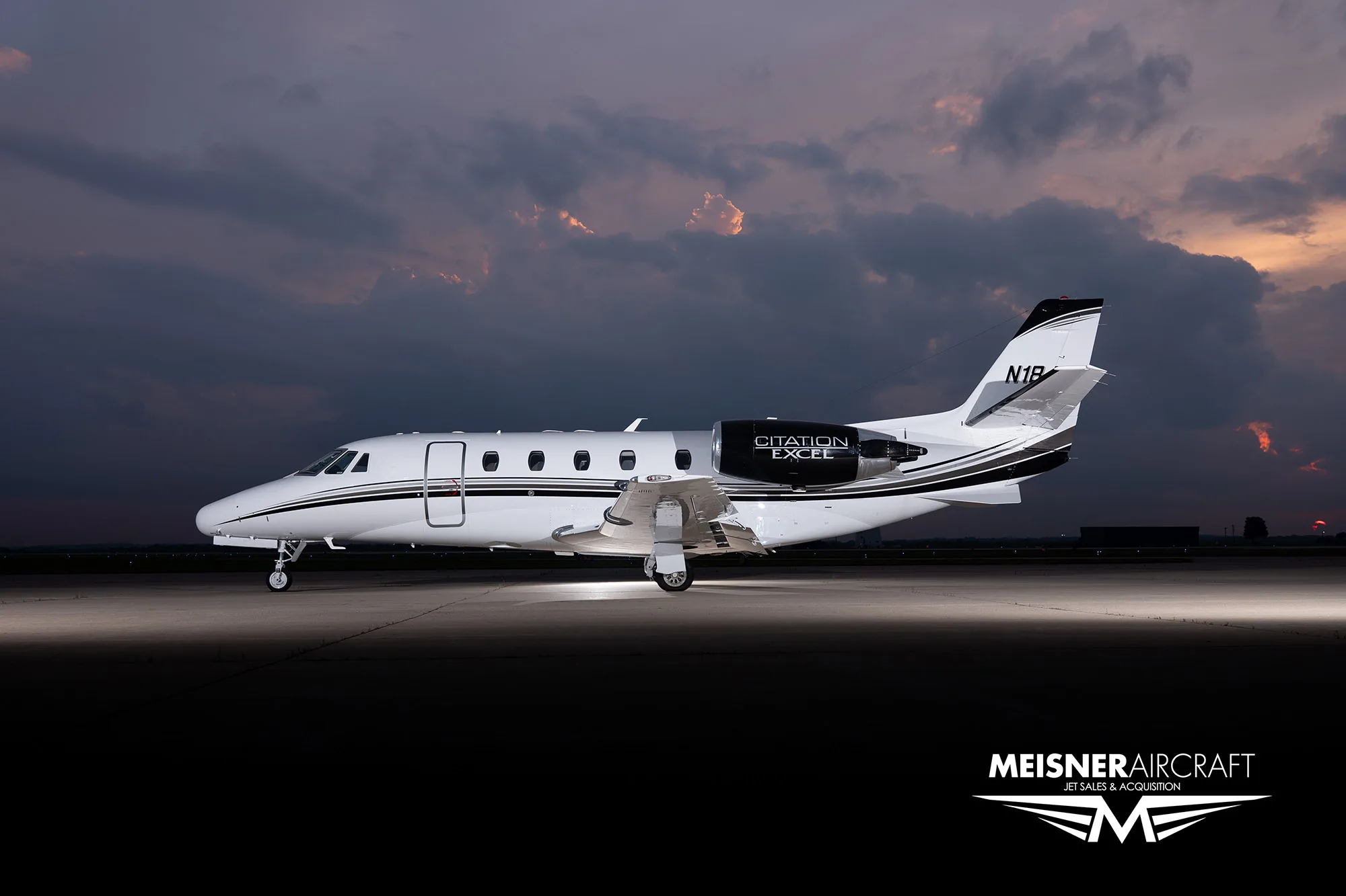 A white Citation Excel business jet with black and white stripes on the tail and engines, parked on a tarmac at dusk.