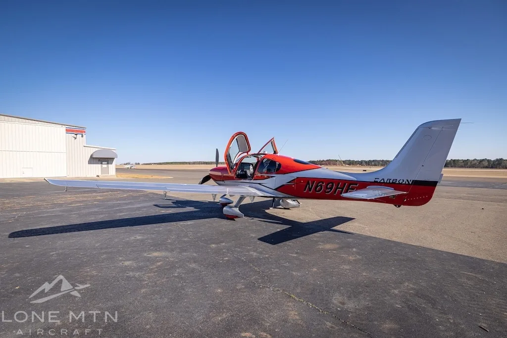 A red and silver Cirrus SR22T aircraft with open doors parked on an asphalt ramp next to a hangar.