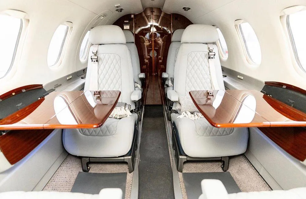 The luxurious interior cabin of the jet, featuring light grey quilted seats and extended wooden tables.