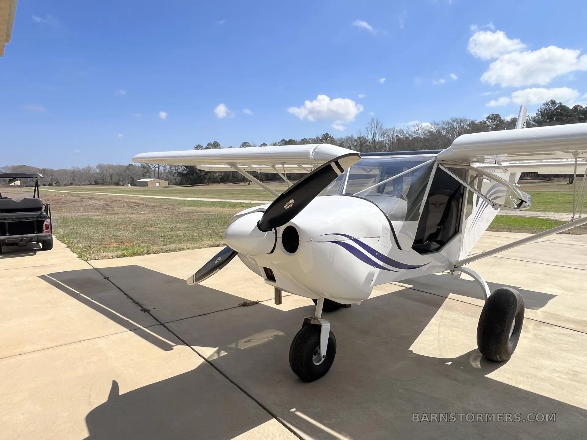 A white high-wing aircraft with blue stripes, parked on a concrete pad with a golf cart nearby.