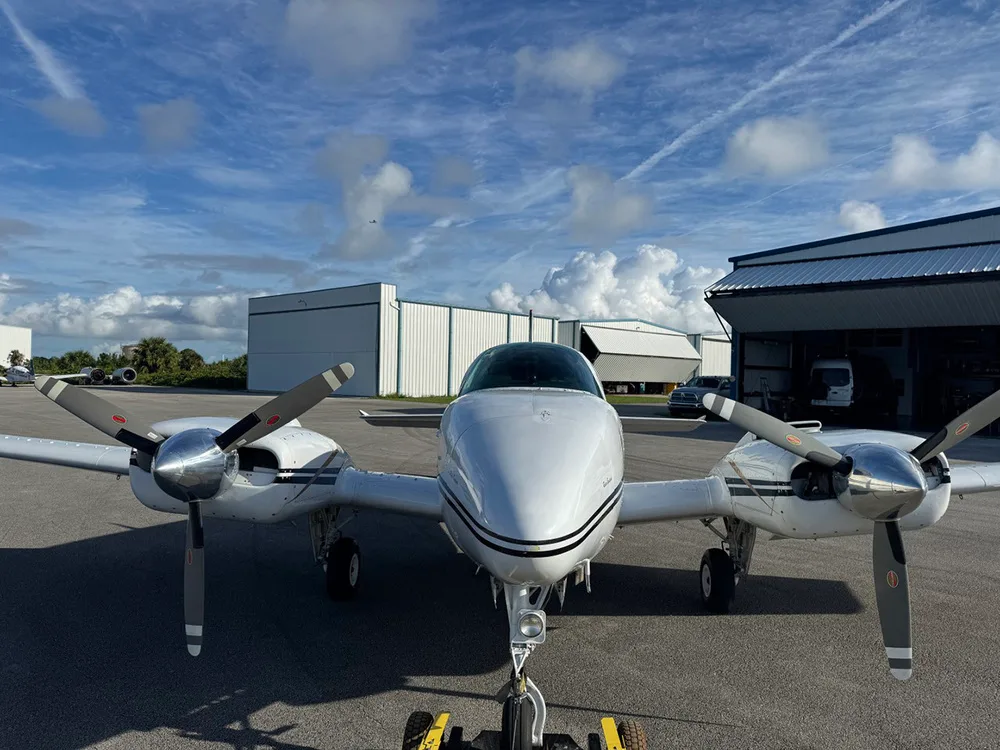 A front view of a white twin-engine Beechcraft Baron aircraft with black stripes, parked on an airfield.