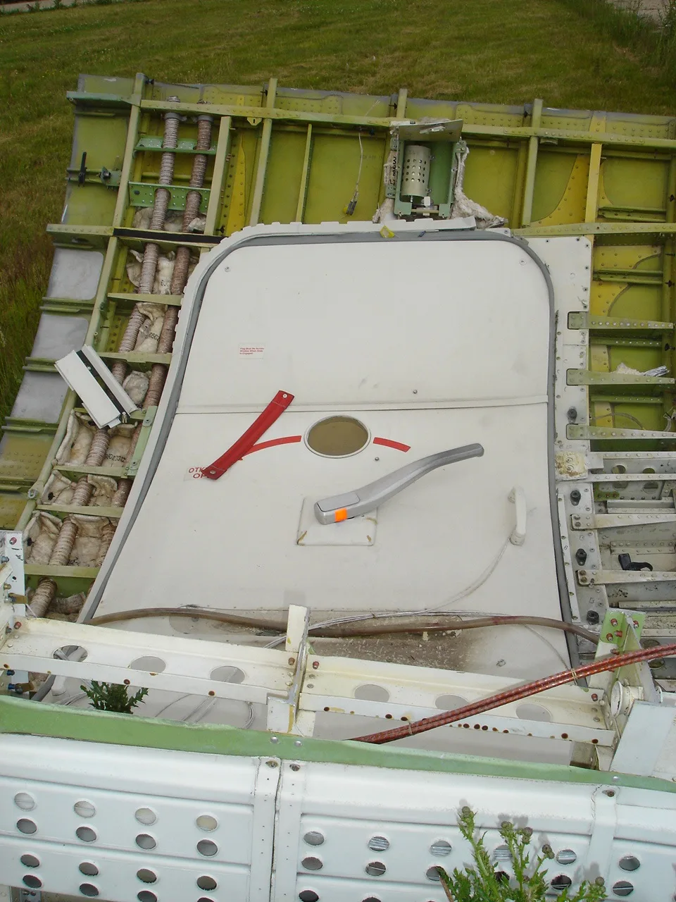A detached aircraft door lying on grass next to exposed fuselage structure.
