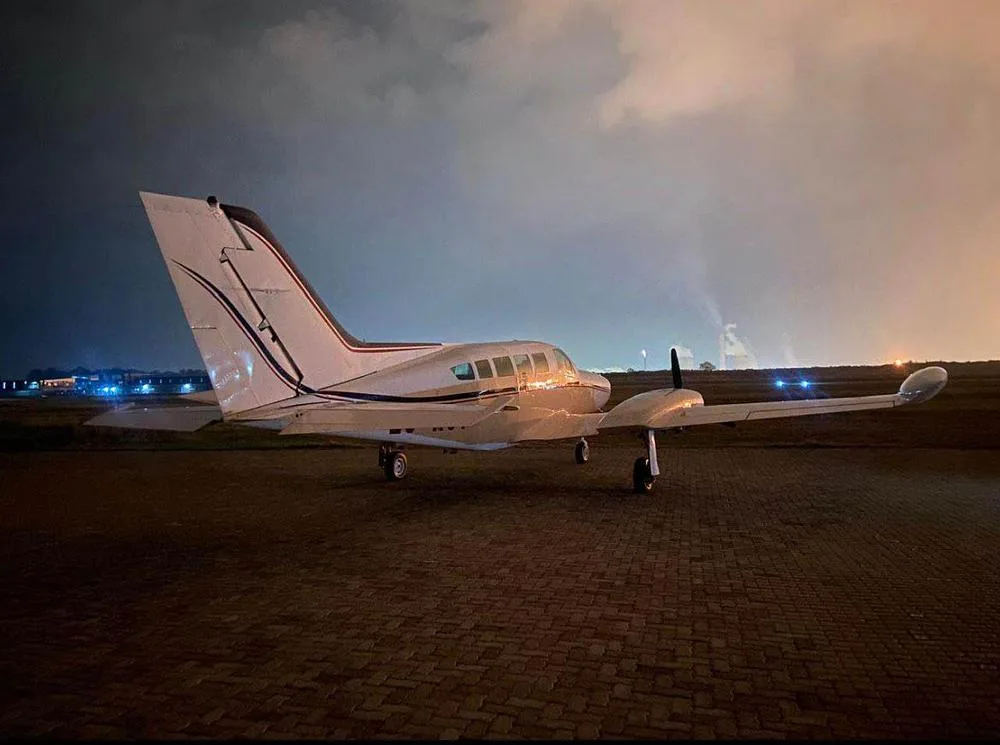 A white twin-engine propeller aircraft with dark blue stripes is parked on a paved surface at night, with distant lights and smoke in the background.