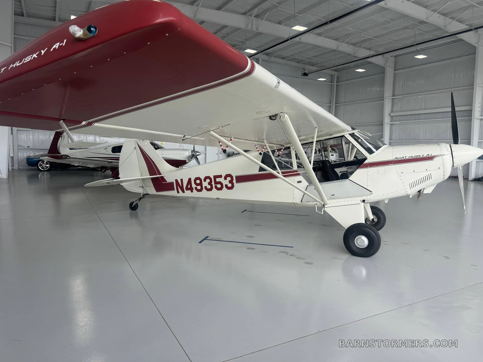 A white and red Aviat Husky A-1 aircraft with registration N49353 is parked in a hangar, viewed from the side.