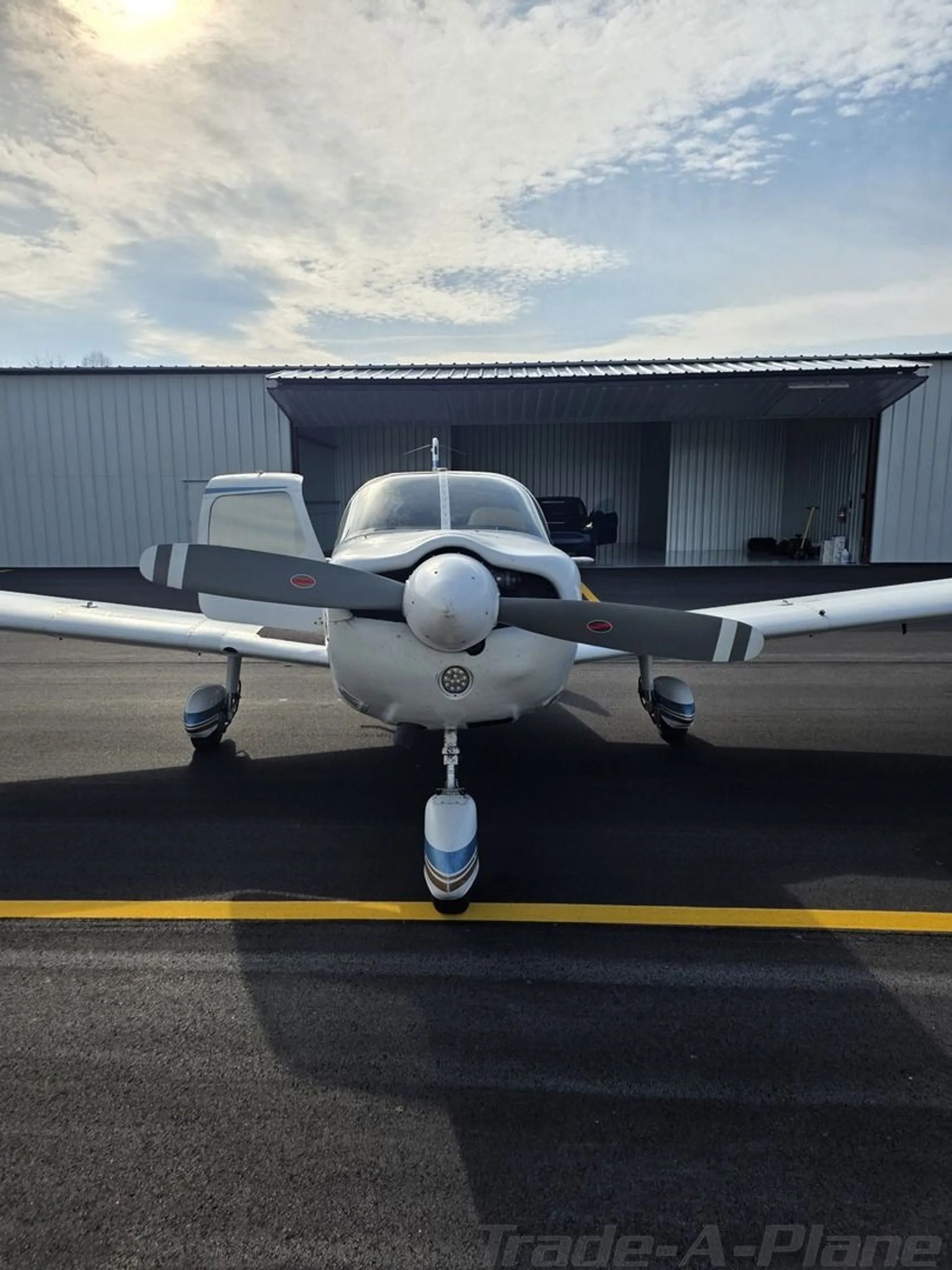 Front view of a white and blue aircraft with a propeller, parked on a tarmac in front of a hangar.
