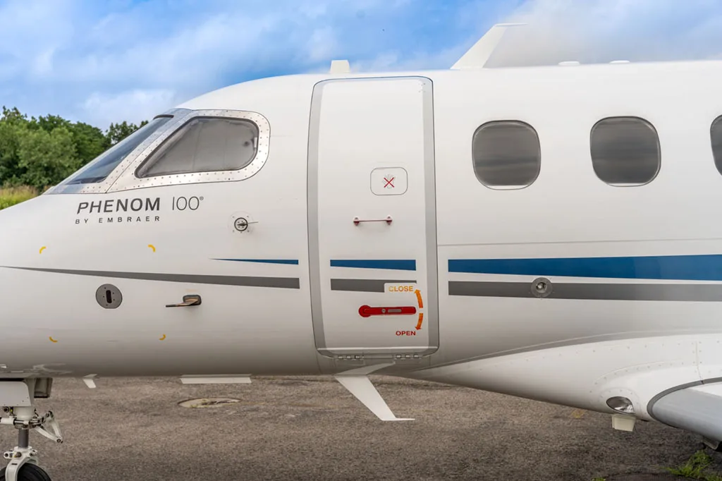 A close-up of the main cabin door and front fuselage of an Embraer Phenom 100, showing the door's open/close instructions.