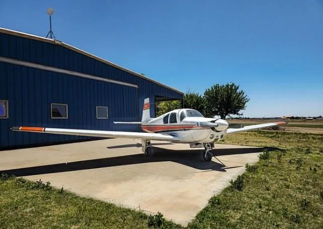 A white and orange Mooney M20 aircraft parked on concrete in front of a blue hangar under a clear sky.