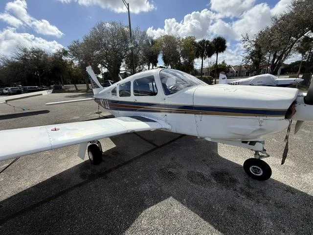 A front-side view of the white Piper Cherokee aircraft, showing its propeller and blue and red stripes.