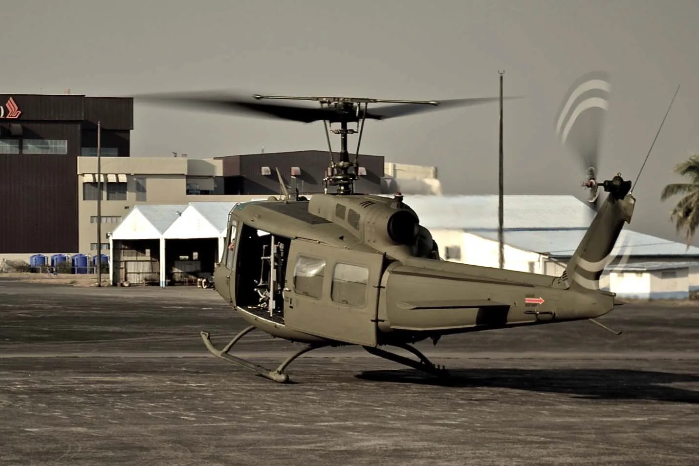 An olive drab utility helicopter, possibly a Bell UH-1 Huey, with spinning rotors sits on a tarmac with buildings in the background.