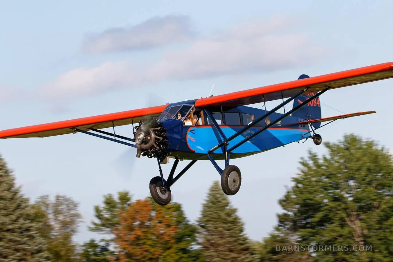 A vintage blue and orange high-wing monoplane, registration NC 9084, flying low over green trees.