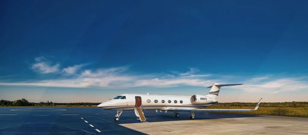 A white Gulfstream G550 private jet with blue and black stripes is parked on a tarmac with its passenger door open and stairs extended, under a clear blue sky.