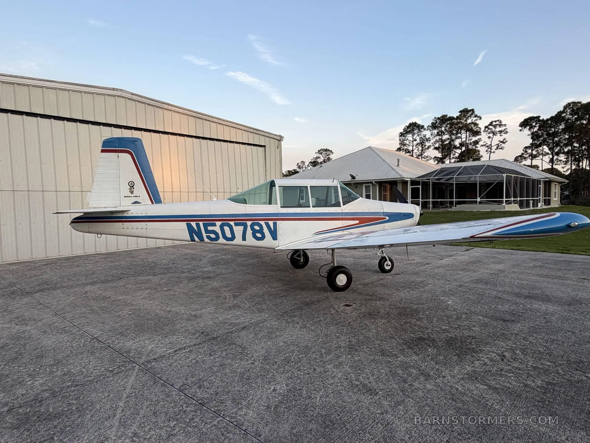 A white and blue single-engine aircraft with registration N5078V, parked on a concrete pad with a hangar and house in the background, viewed from the left side.