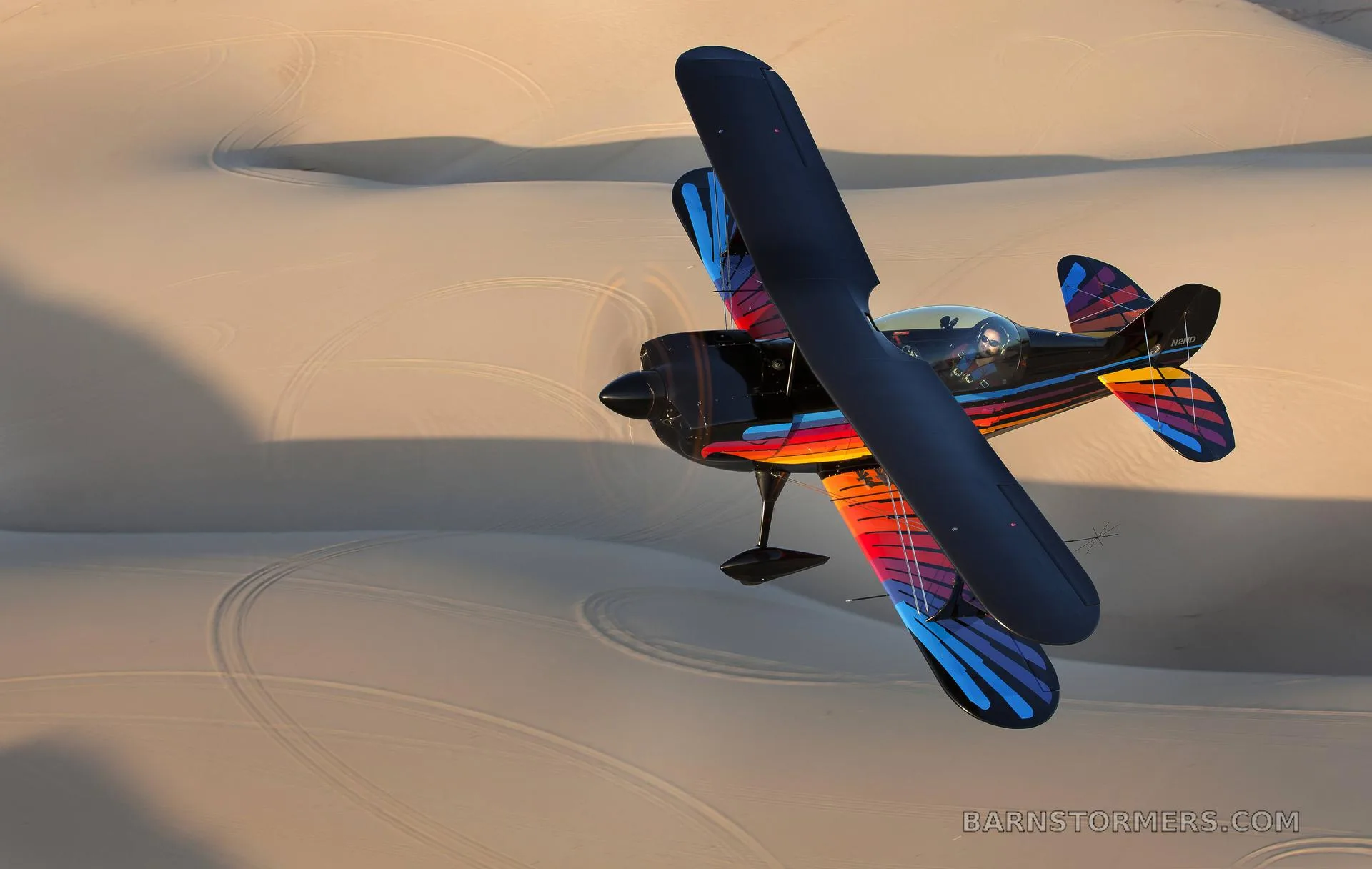 A black biplane with a vibrant red, orange, and blue paint scheme on its wings and tail, flying over vast sand dunes.
