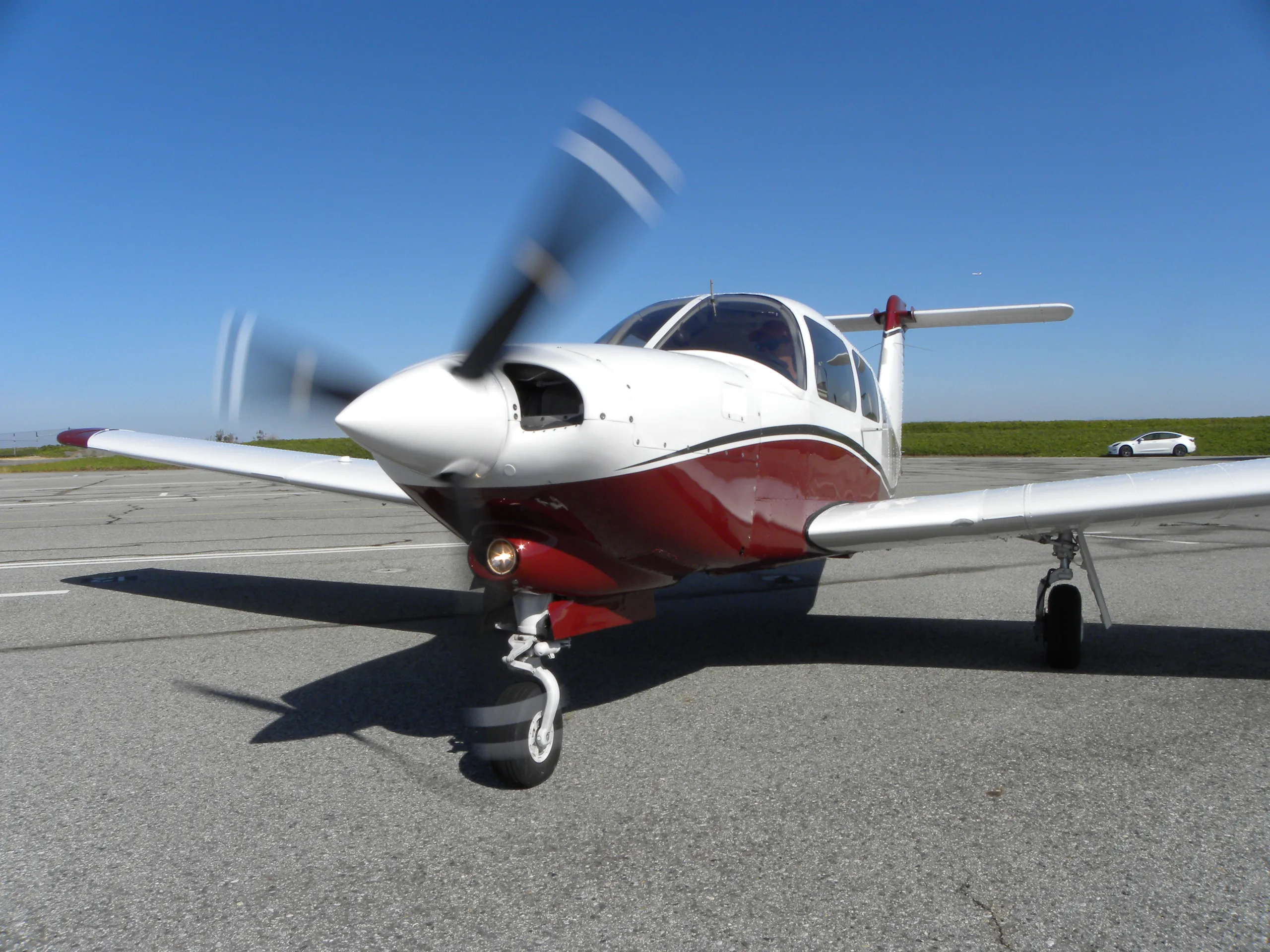 A white and red single-engine propeller aircraft with a spinning propeller sits on an asphalt tarmac under a clear blue sky.