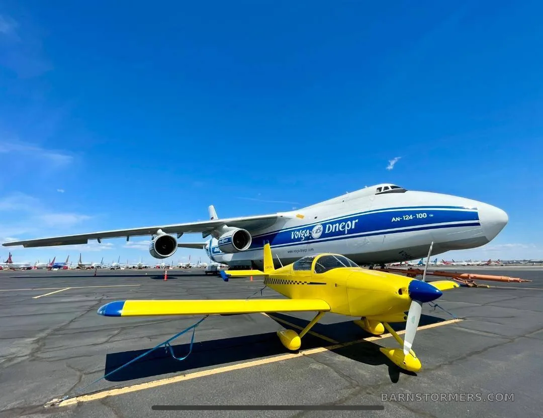 A small yellow aerobatic aircraft with a blue propeller tip is parked on an airfield, with a large white and blue Volga-Dnepr AN-124 cargo plane in the background.