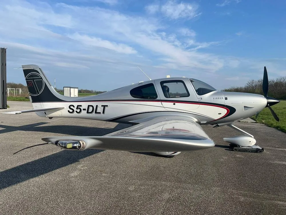 A silver and white Cirrus SR22T aircraft with registration S5-DLT parked on an asphalt surface under a partly cloudy sky.