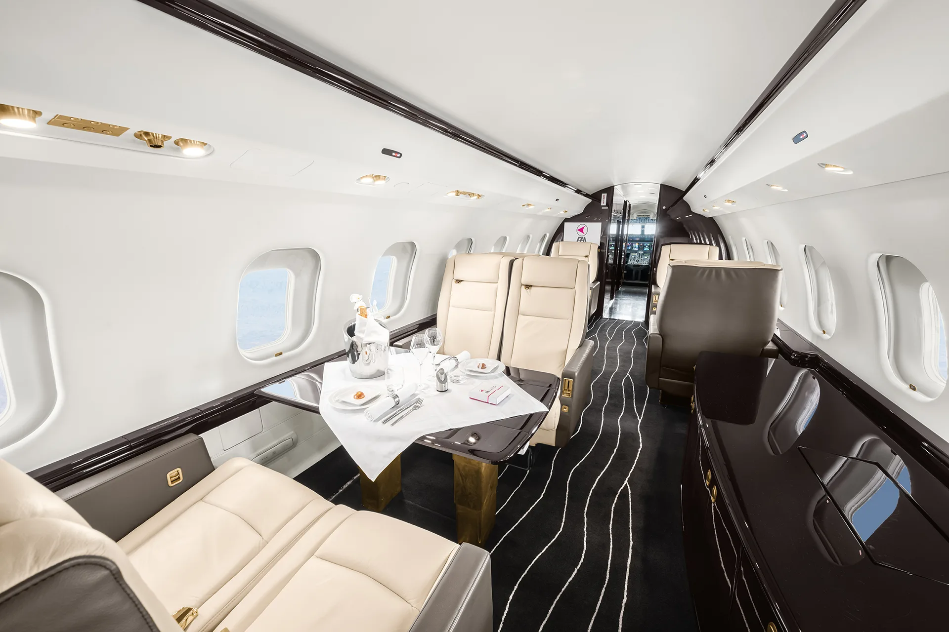 A private jet cabin interior featuring a set dining table with champagne and place settings, cream seats, and dark wood finishes.