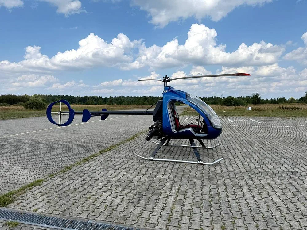 A blue single-seat helicopter with a clear bubble canopy is parked on a paved surface under a blue sky with white clouds.