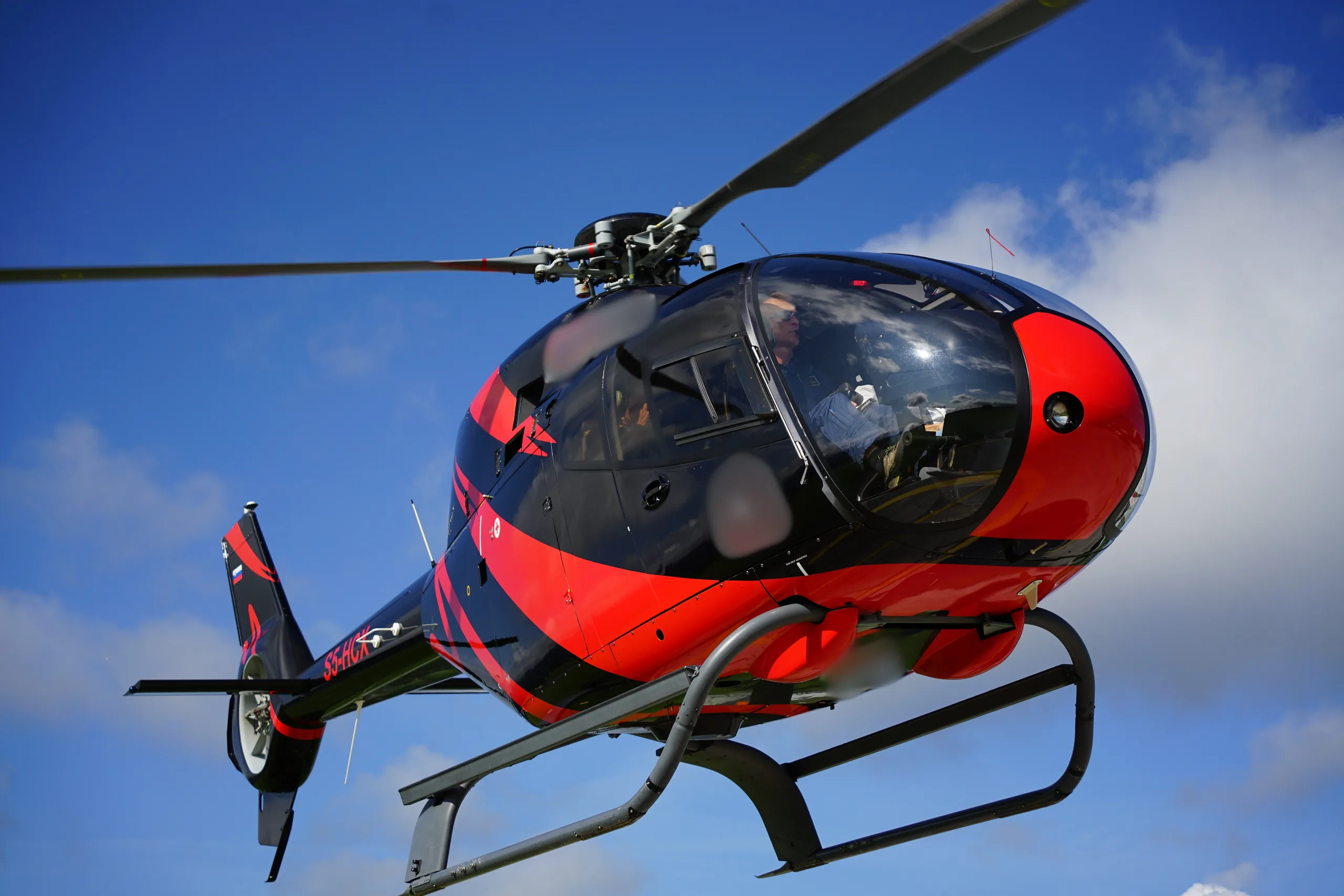 A black and red helicopter is shown from a low angle, with a blue sky in the background.