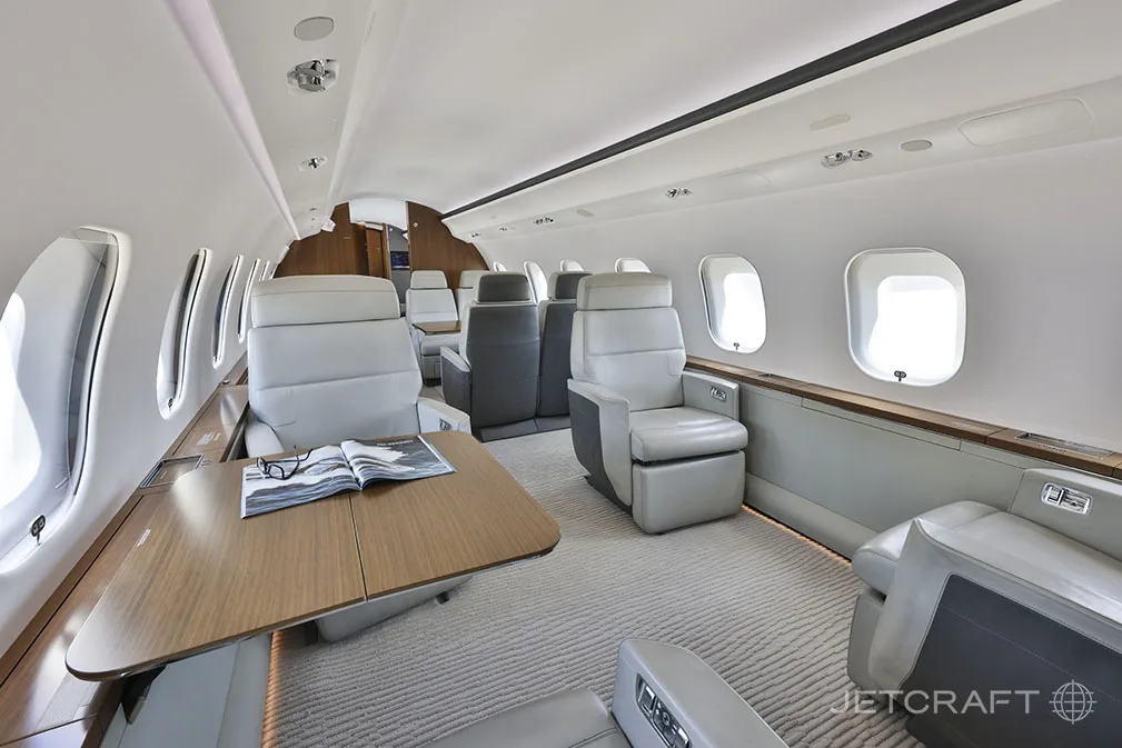 A view inside the Bombardier Global 6000 cabin, showing light grey leather seats, a wooden table with a magazine, and large windows.