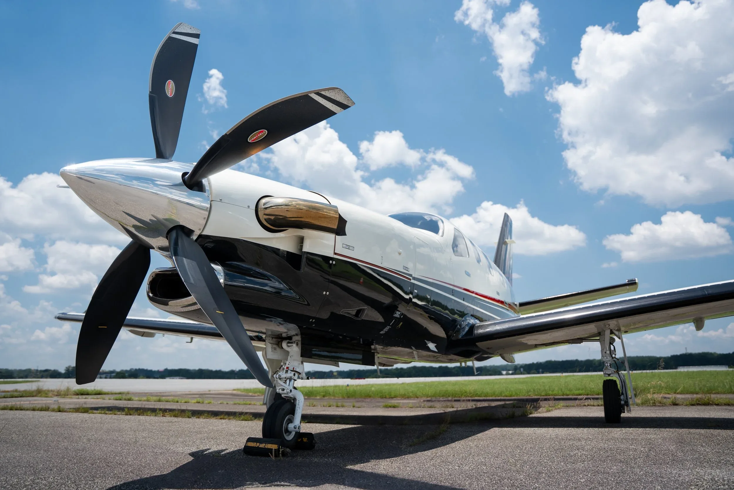 A low-angle front-quarter view of a white and black TBM 960 aircraft with its propeller, parked on an airport tarmac under a blue sky.