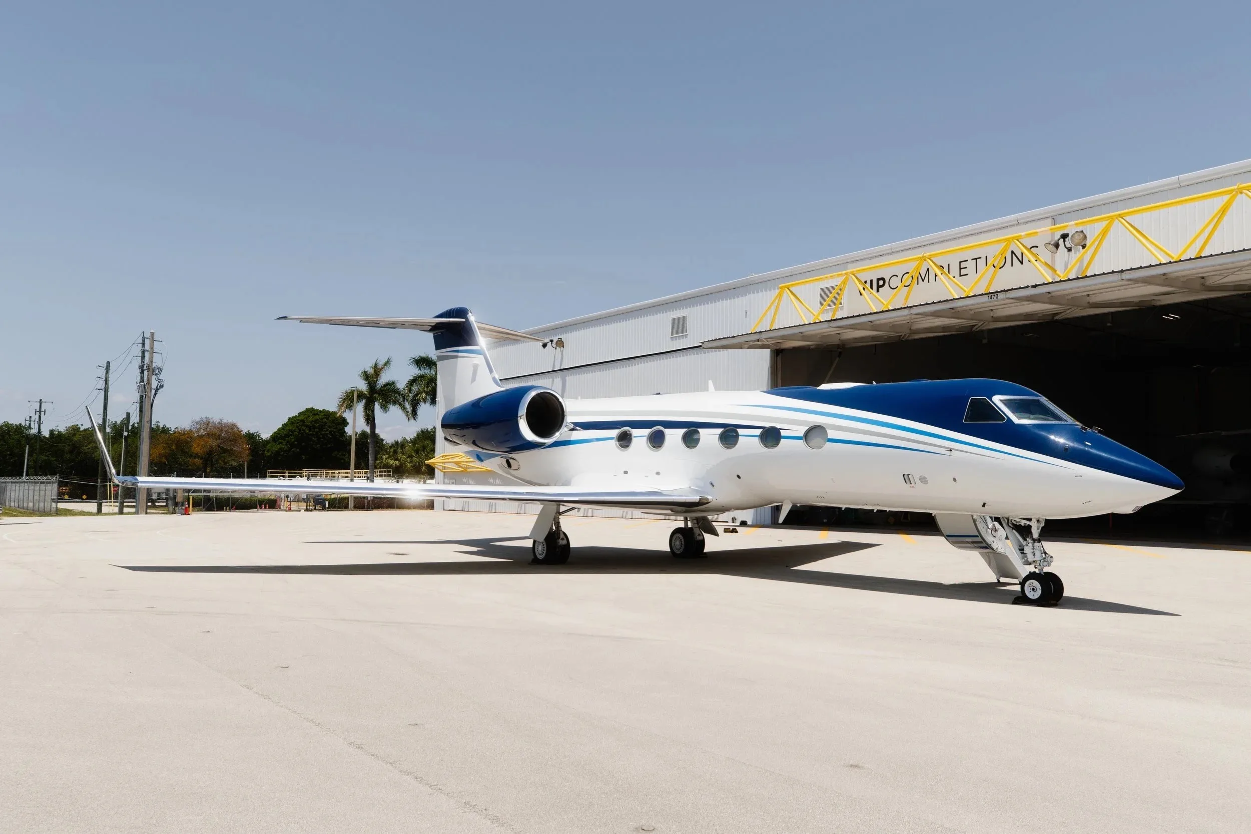 A Gulfstream G550 business jet with a white and blue paint scheme parked on an airfield next to a hangar.