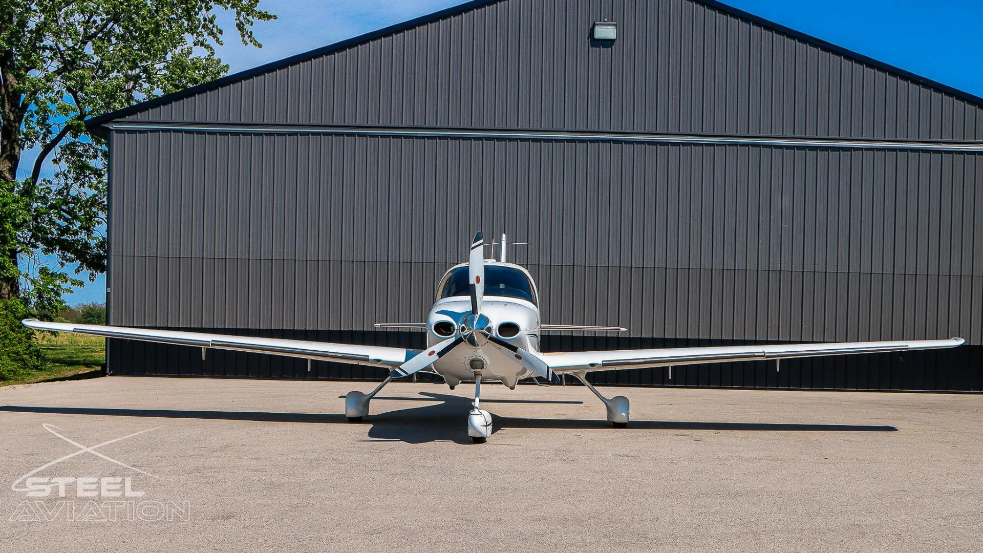 Front view of a white Cirrus SR22 aircraft with black and grey stripes, parked in front of a grey hangar.
