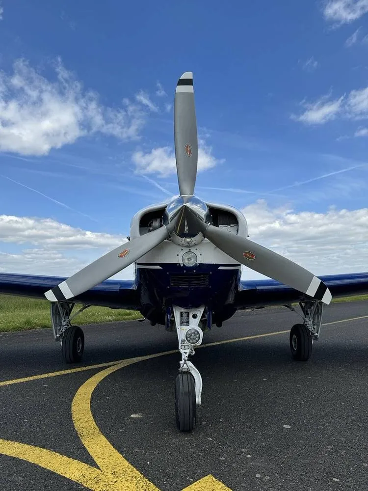 A head-on view of the propeller and nose of a white and blue Beechcraft Bonanza aircraft on an airfield.