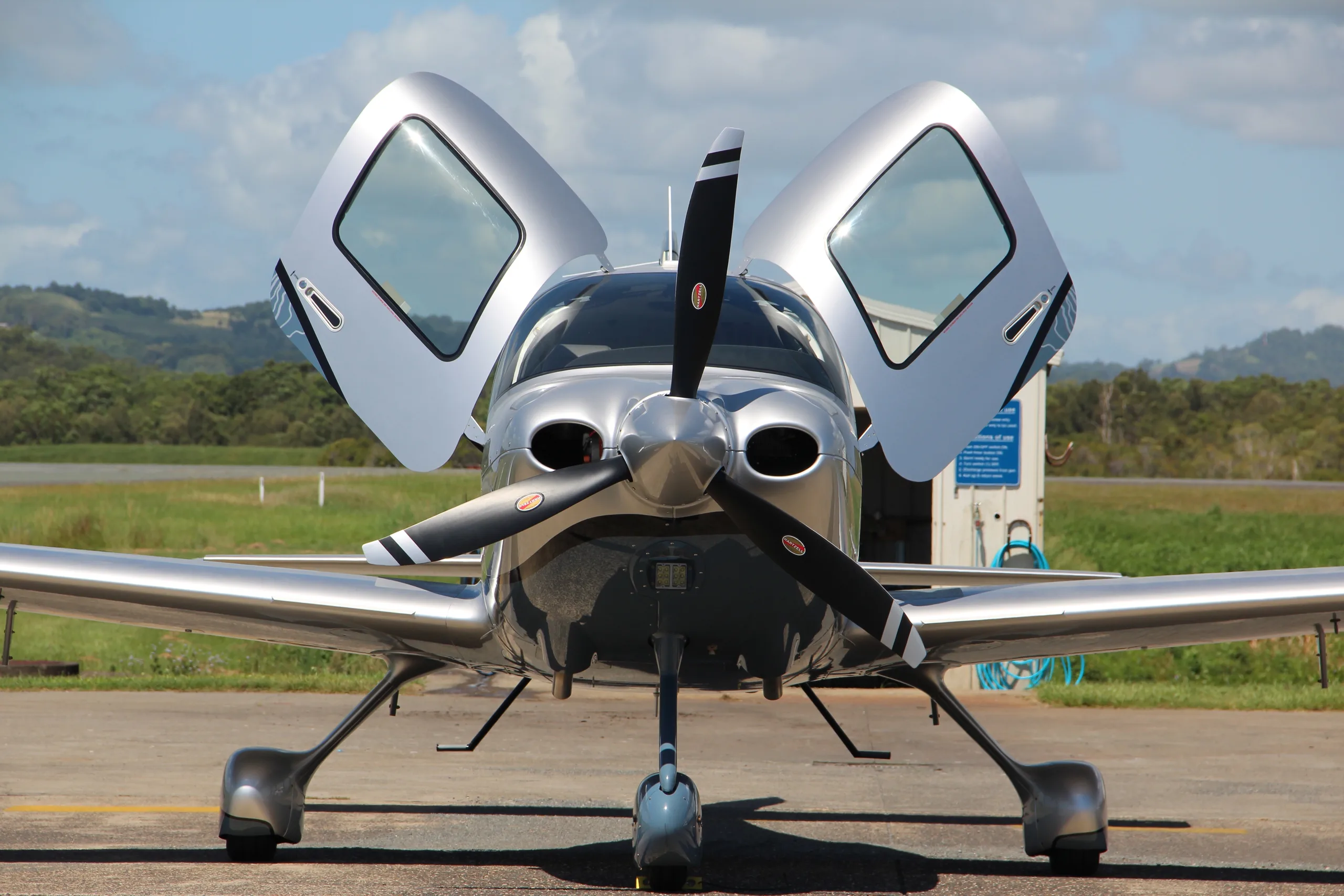 A silver single-engine propeller aircraft with open gull-wing doors parked on an airfield, with green hills in the background.